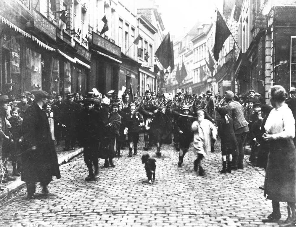 Photographie en noir et blanc – Le 42e bataillon, jouant de la cornemuse et défilant en fendant une foule de citoyens français dans les rues de Mons.